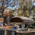 Ein Ooni Koda 2 Gas-Pizzaofen von Ooni steht auf einem Metalltisch, während ein verschwommener Mann und ein Kind in einem sonnenbeschienenen, von Bäumen gesäumten Hinterhof gemeinsam neapolitanische Pizza backen.