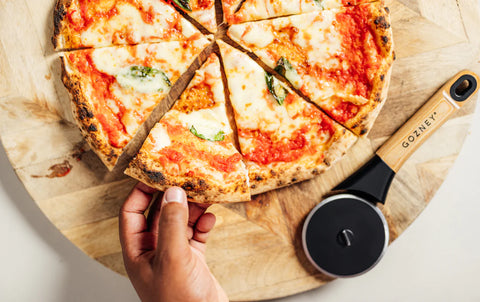 Eine Hand nimmt ein Stück Pizza von einem hölzernen Pizzabrett. Die Pizza hat einen dünnen Boden, belegt mit geschmolzenem Käse, Tomatensauce und Basilikumblättern. Neben der Pizza liegt ein Gozney Pizzaschneider von Gozney mit einem ergonomischen Griff, der professionelle Schneidequalität verspricht.