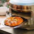 Eine heiße 12-Zoll-Gemüse-Käse-Pizza wird mit einem Metall-Pizzaschieber aus dem Solo Stove Pi Pizzaofen 12 von Solo Stove geholt, während Dampf im Hintergrund auf die hohen Backtemperaturen hinweist.