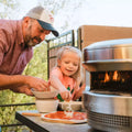Ein Mann hilft einem jungen Mädchen, den 12-Zoll-Pizzateig im Freien mit Käse zu bestreuen. Ein Solo Stove Pi Pizzaofen 12 von Solo Stove, mit Flammen im Inneren, steht auf dem Tisch inmitten von üppigem Grün im Hintergrund.