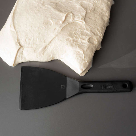 Eine Portion rohen Teigs liegt auf einer glatten, grauen Fläche. Unter dem Teig liegt ein schwarzer, flexibler Teigspachtel mit Griff, waagerecht platziert. Die Fläche um Teig und Spachtel ist sauber und aufgeräumt – perfekt für Pizza-Enthusiasten.