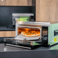 Der Pizzaofen Effeuno P134H 509°C Evolution mit Biscotto Pizzastein backt eine Pizza mit sichtbaren Belägen wie Basilikum und Mozzarella und erreicht damit neapolitanische Pizzaqualität. Die Ofentür ist geöffnet, die Pizza liegt leicht herausgezogen auf einem Blech. Im Hintergrund ist eine moderne Küchenkulisse mit einem weiteren Kompaktgerät zu sehen.