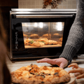 Eine Person mit grauem Ärmel ist von der Seite zu sehen und hält eine Platte mit einer frisch gebackenen neapolitanischen Pizza. Im Hintergrund wird eine weitere Pizza in einem Effeuno Pizzaofen Effeuno P134HA 509°C Evolution mit Biscotto Pizzastein gebacken. Ein Paar junger Hände streckt sich nach der Pizza auf der Platte aus.