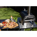 Ein Ooni Karu 2 Outdoor-Pizzaofen von Ooni steht auf einem Metalltisch im Freien, neben einer Margherita-Pizza auf einer hölzernen Schale, mit Schalen mit Belag, frischem Basilikum und Gewürzen in der Nähe in der Sonne.