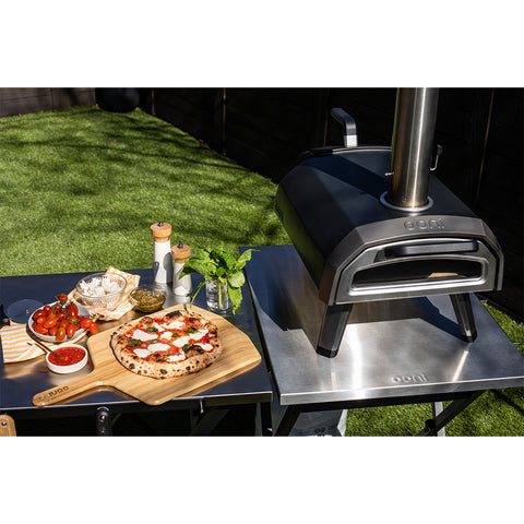 Ein Ooni Karu 2 Outdoor-Pizzaofen von Ooni steht auf einem Metalltisch im Freien, neben einer Margherita-Pizza auf einer hölzernen Schale, mit Schalen mit Belag, frischem Basilikum und Gewürzen in der Nähe in der Sonne.