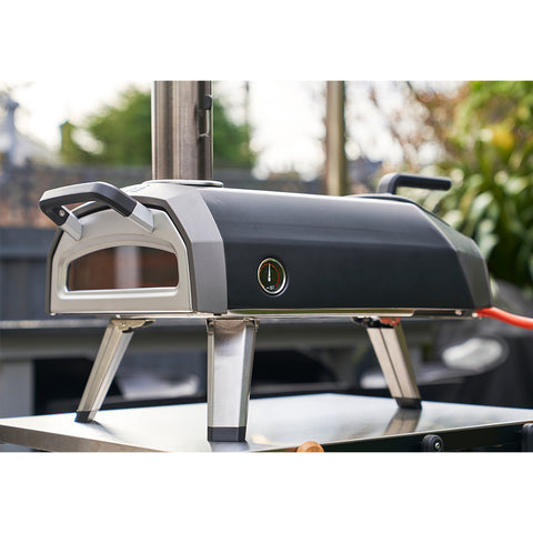 Ein schwarz-silberner Ooni Karu 2 Outdoor-Pizzaofen von Ooni mit seitlichem Thermometer steht tragbar auf einem Metalltisch mit verschwommenem Grün und einem Zaun im Hintergrund.