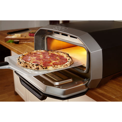 Eine neapolitanische Pizza mit Peperoni wird mithilfe einer Pizzaschaufel aus einem offenen Ooni Volt 12 Elektro Pizzaofen geholt. Der Ofen steht auf einer Holzarbeitsplatte, im Hintergrund sind einige Zutaten zu sehen, die praktischerweise über eine nahe gelegene 230 V-Steckdose mit Strom versorgt werden.