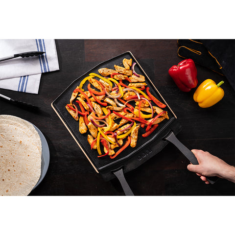 Eine Hand hält den Griff einer doppelseitigen Grillpfanne von Ooni, die mit gekochten Hähnchenstreifen, roten und gelben Paprikaschoten und roten Zwiebeln gefüllt ist. In der Nähe liegen eine Zange, ein weiß-blau gestreiftes Handtuch, frische Paprikaschoten und ein Teller Tortillas auf dem dunklen Holztisch.
