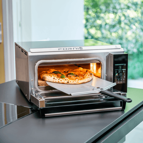 Der Pizzaofen Effeuno P134H 509°C Evolution mit Biscotto Pizzastein präsentiert bei geöffneter Tür eine frisch gebackene Pizza, die fachmännisch mit einer Pizzaschaufel herausgezogen wird. Die neapolitanische Pizzaqualität zeigt sich in der goldenen Kruste, dem geschmolzenen Käse und den sichtbaren Basilikumblättern. Dieser Premium-Ofen von Effeuno befindet sich in einer modernen Küche, durch deren Fenster man Grünpflanzen sehen kann.