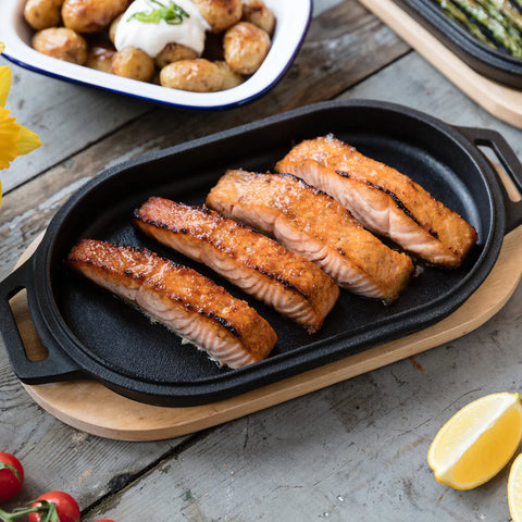 Eine Ooni Servierpfanne aus Gusseisen mit vier gegrillten Lachsfilets ruht auf einem Holzbrett. Der Lachs hat eine goldbraune Außenseite. Im Hintergrund befindet sich ein Gericht aus Bratkartoffeln mit Sauerrahm und Schnittlauch sowie Spargel. Eine Zitronenscheibe und Tomaten stehen in der Nähe und ergänzen dieses Ooni-Juwel auf seinem Holzboden.
