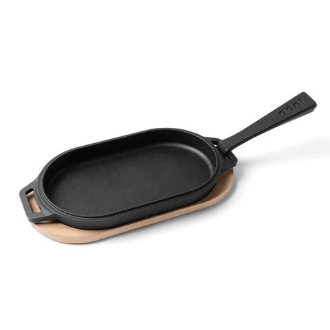Eine schwarze Grillpfanne aus Gusseisen, bekannt als Ooni Servierpfanne aus Gusseisen, mit Griff steht auf einem Holzboden-Servierbrett. Die oval geformte Pfanne verfügt auf der gegenüberliegenden Seite über einen zusätzlichen kleinen Griff. Das Holzbrett ist so gestaltet, dass es perfekt auf den Boden der Ooni-Pfanne passt.