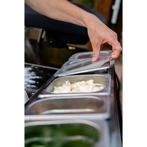 In einer geschäftigen Küche hebt jemand den Deckel von einem Behälter voller Teig oder Nudeln und bereitet eine Pizzaparty vor. Auf der Arbeitsplatte präsentiert eine Ooni Pizza Topping Station von Ooni verschiedene Zutaten und Beläge. Der leicht unscharfe Hintergrund verstärkt den Fokus auf diesen sorgfältigen Zubereitungsprozess.
