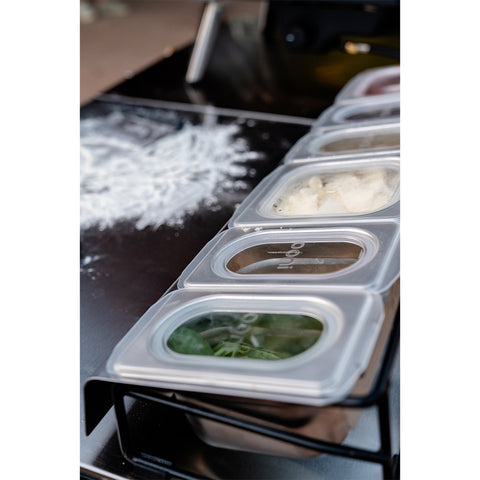 Eine Reihe transparenter Behälter mit Deckel, Teil der Ooni Pizza Topping Station von Ooni, ist auf einer schwarzen Oberfläche angeordnet, die mit etwas bestreut ist, das wie Mehl aussieht. Die Behälter sind ideal für die Vorbereitung einer Pizza-Party und enthalten eine Vielzahl von Zutaten wie Kräuter, Käse und möglicherweise Teig.