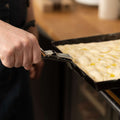 Eine Hand hält die FUMOSA Zange für Pizzablech von Fumosa und zieht damit ein mit Olivenöl bestrichenes Teigblech aus dem Ofen. Im Hintergrund sind eine hölzerne Arbeitsplatte und eine Kücheneinrichtung zu sehen, die für eine sichere Entnahme sorgt.