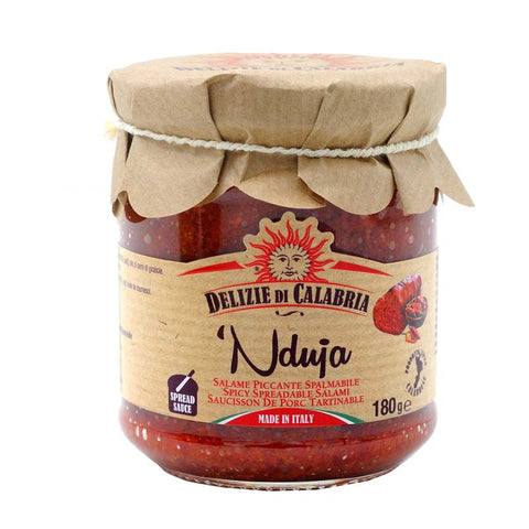 Ein Glas „Delizie di Calabria Nduja Calabrese im Glas (Streichsalami),“ eine würzige streichfähige Schweinefleischpastete aus Kalabrien, Italien. Das Glas ist mit einem braunen Papierdeckel verschlossen, der mit einer Schnur zusammengebunden ist. Das Etikett gibt an, dass es sich um eine 180 g (6,35 oz) Streichsalami handelt. Das Design ist in den Farben Rot und Weiß mit Chili-Grafiken gestaltet.