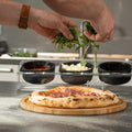Eine Person belegt eine frisch gebackene Pizza in einer FUMOSA Pizzastation POMPEY mit Blattgemüse. Die Pizza liegt auf einem Holzbrett, dahinter stehen Schüsseln mit verschiedenen Zutaten wie Soße und Käse. Die Hände der Person, geschmückt mit einer braunen Uhr, handhaben den Belag gekonnt.