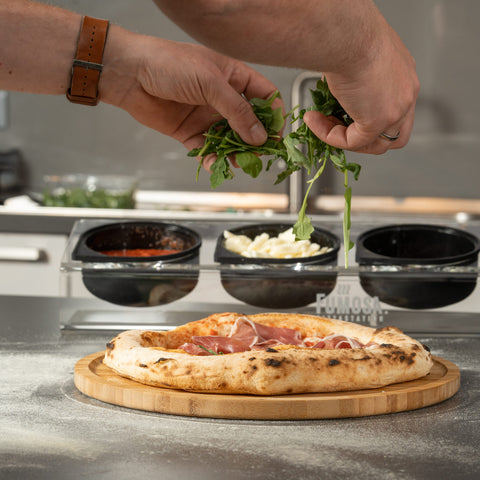 Eine Person belegt eine frisch gebackene Pizza in einer FUMOSA Pizzastation POMPEY mit Blattgemüse. Die Pizza liegt auf einem Holzbrett, dahinter stehen Schüsseln mit verschiedenen Zutaten wie Soße und Käse. Die Hände der Person, geschmückt mit einer braunen Uhr, handhaben den Belag gekonnt.
