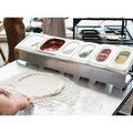Eine Person formt einen Pizzateig auf einer bemehlten Fläche. Vor ihr steht eine Ooni Pizza Topping Station von Ooni mit durchsichtigen Deckeln mit der Aufschrift „Ooni“, die Zutaten wie Tomatensoße, Käse und verschiedene Beläge enthalten. Im leicht verschwommenen Hintergrund bereiten sie sich auf eine lustige Pizza-Party vor.