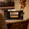 Ein Pizzaofen Effeuno P134HA 509°C Evolution mit Biscotto Pizzastein steht auf einer Küchenarbeitsplatte. Der Ofen hat ein digitales Bedienfeld und eine große Tür. Die Küche ist mit warmen Schränken, gefliester Küchenrückwand und herbstlichen Dekorationen an der Wand ausgestattet. Im Vordergrund ist ein Teil eines Tisches mit Stuhl zu sehen, perfekt, um selbstgemachtes Biscotto zu genießen.