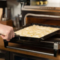 Man sieht eine Hand, die eine Pfanne mit Teig oder Fladenbrot hält und diese in einen offenen Tischbackofen aus hochwertigem Edelstahl schiebt. Der Markenname des Ofens, Forno, ist auf der Tür zu sehen. Daneben steht eine FUMOSA Zange für Pizzablech der Marke Fumosa auf der Küchentheke, im Hintergrund sind außerdem verschiedene Utensilien und Pizzableche zu sehen.
