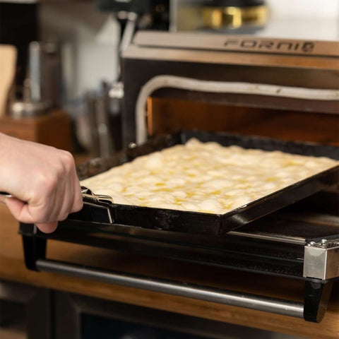 Man sieht eine Hand, die eine Pfanne mit Teig oder Fladenbrot hält und diese in einen offenen Tischbackofen aus hochwertigem Edelstahl schiebt. Der Markenname des Ofens, Forno, ist auf der Tür zu sehen. Daneben steht eine FUMOSA Zange für Pizzablech der Marke Fumosa auf der Küchentheke, im Hintergrund sind außerdem verschiedene Utensilien und Pizzableche zu sehen.