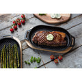 Ein gegrilltes Steak mit einer Scheibe Kräuterbutter liegt auf einer Ooni Grillpfanne aus Gusseisen und weist perfekte Grillstreifen auf. Daneben hält eine weitere Grillpfanne aus Gusseisen mit abnehmbarem Griff grüne Spargelstangen. Frische Kirschtomaten und Petersilie sind auf der rustikalen Holzoberfläche verstreut, zusammen mit einem Holzbrett, auf dem noch mehr Kräuterbutter liegt.
