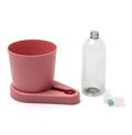 Der greevi Kräuterheld selbstbewässernder Kräuter-Pflanztopf (GREEVI) von Greevi, abgebildet in Rosa mit passendem Sockel, sitzt neben einer klaren Flasche, einer weißen Kappe und einer grünen Kugel - perfekt für die einfache Kräuterbewässerung.