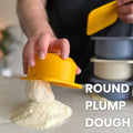 Eine Person hält ein Babadoh Silikon BIG Pizzateig und Zutatenbehälter 3er-Set, das auf einem Berg mit Mehl bestreuter Pizzateigkugeln ruht. Der Text „ROUND PLUMP DOUGH“ ist in klarer, weißer Schrift auf der rechten Seite des Bildes deutlich zu sehen. Im Hintergrund sind mehrere Schüsseln gestapelt, die die Szenerie für ein echtes Pizza-Abenteuer schaffen.