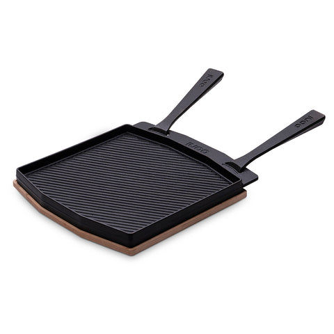 Abbildung einer doppelseitigen Grillpfanne von Ooni, einer schwarzen, rechteckigen Gusseisenpfanne mit zwei Griffen und einer gerippten Struktur auf der Kochfläche, die zum Grillen konzipiert ist. Die Pfanne steht auf einem passenden hitzebeständigen Untersetzer.