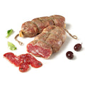 Zwei große Stücke Madeo Soppressata Salami BIO (Mild) liegen mit Schnüren zusammengebunden auf einer weißen Fläche, davor sind drei Scheiben der Salami aufgeschnitten und ausgestellt. Neben der Salami liegen zwei schwarze Oliven und einige grüne Blätter.