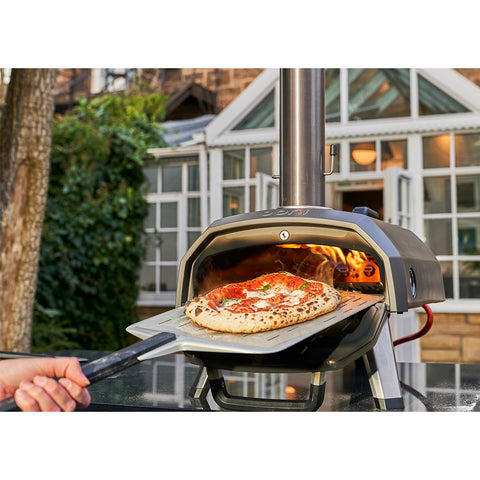 Eine Hand schiebt eine frisch gebackene Pizza mit Tomaten und Kräutern aus dem Ooni Karu 2 Outdoor-Pizzaofen von Ooni, während im Hintergrund ein Haus und Grünflächen zu sehen sind.