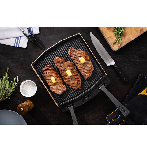 Drei gegrillte Steaks mit Butterstückchen brutzeln auf einer schwarzen doppelseitigen Grillpfanne von Ooni. Die Pfanne steht auf einer dunklen Oberfläche, daneben liegen Rosmarinzweige, ein Messer, eine Schüssel mit grobem Salz, eine Gabel und eine Pfeffermühle. Ein Schneidebrett mit Rosmarin und ein Küchentuch sind ebenfalls zu sehen.